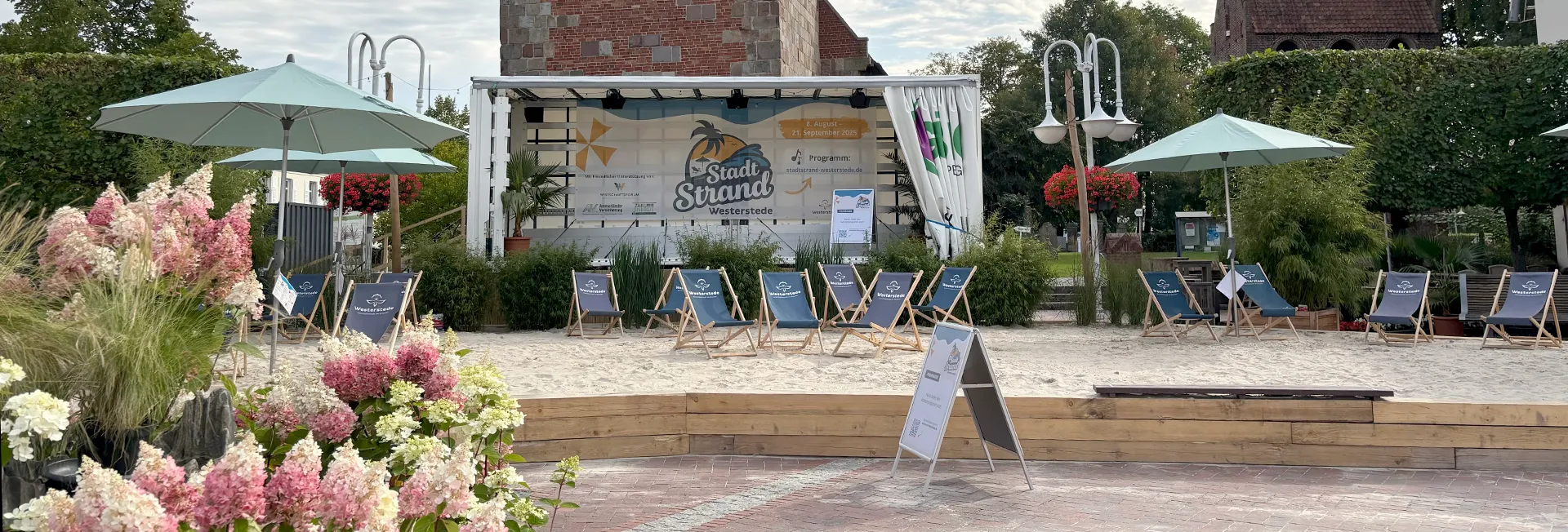 Außengelände mit Sandstrand, Liegestühlen, Sonnenschirmen und einer weißen Event-Bühne