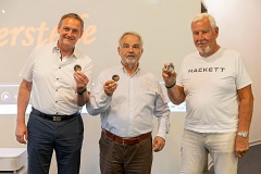 2026-06-21_Veteranencafe_Coins_97A2886.png Drei Männer halten kleine runde Abzeichen in ihren Händen vor einer weißen Wand.