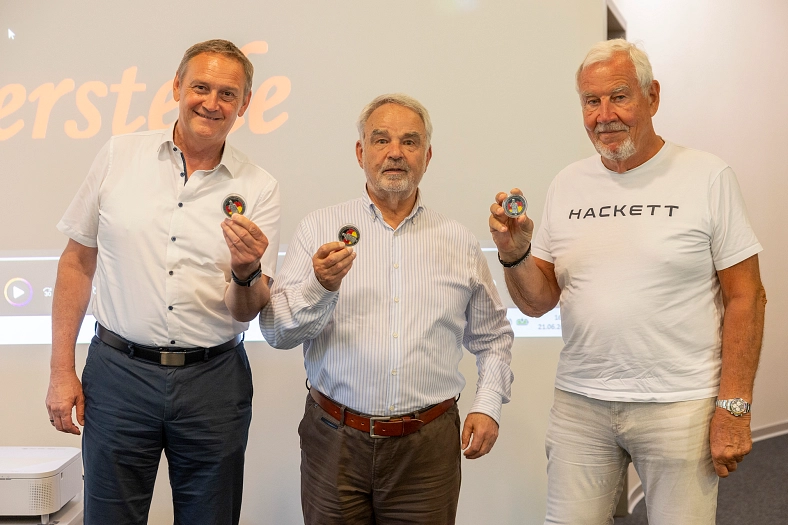 Drei Männer halten kleine runde Abzeichen in ihren Händen vor einer weißen Wand.