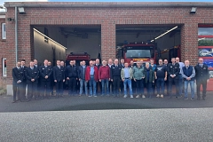 2026-06_Feuerwehrgeraeteschau.jpg Gruppe von Feuerwehrleuten und Ehrengästen vor einem Feuerwehrfahrzeug in einer Ziegelgarage.